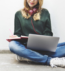 Dame met laptop op schoot en koptelefoon om haar nek die schrijft in een kladblok