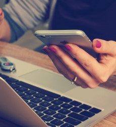 Foto van een vrouw die een mobiele telefoon vasthoudt en achter haar laptop zit.