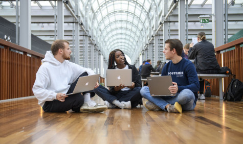Drie studenten zitten op de grond en zijn in gesprek. Ze werken op hun laptops.