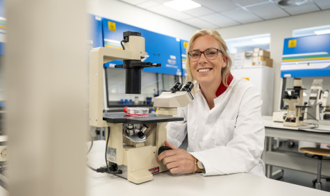 Anna Posthumus zit achter een microscoop in een lab, ze kijkt lachend de camera in. 