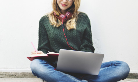 Dame met laptop op schoot en koptelefoon om haar nek die schrijft in een kladblok