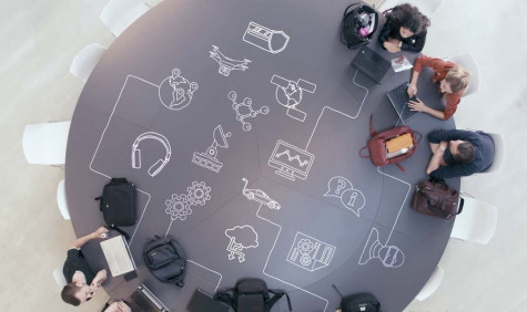 Studenten werkend aan een ronde tafel, van bovenaf bekeken. Op de tafel staan illustraties die de verbinding tussen ICT, Onderwijs en Onderzoek verbeelden