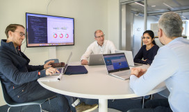 Vier mensen aan een ronde tafel met laptops en presentatiescherm