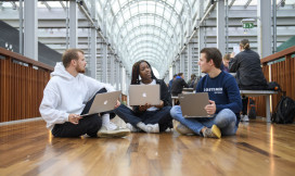 Drie studenten zitten op de grond en zijn in gesprek. Ze werken op hun laptops.