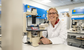 Anna Posthumus zit achter een microscoop in een lab, ze kijkt lachend de camera in. 