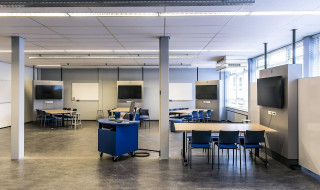 Deactive learing classroom is een collaboratieve onderwijsruimte, waarbij de docent opereert vanuit het midden van de zaal en acht groepen van zes studenten de beschikking hebben over een zit-sta tafel, scherm en whiteboard