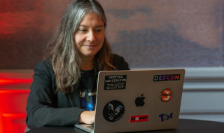 Melanie Rieback werkt op haar laptop waarop stickers zitten van o.a. Defcon en een sticker waarop staat 'Venture capitalism destroys everything you hold dear and makes you pay for the privilege to watch'