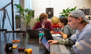 Drie studenten zitten aan een tafel, twee zijn in gesprek en de ander werkt op zijn laptop.