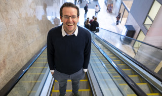 Tom Emery staat op een roltrap en kijkt lachend de camera in 