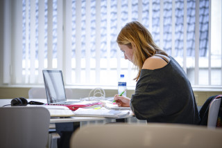 Studente aan het werk achter computer met veel documenten en aantekeningen voor zich