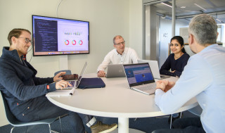 Vier mensen aan een ronde tafel met laptops en presentatiescherm