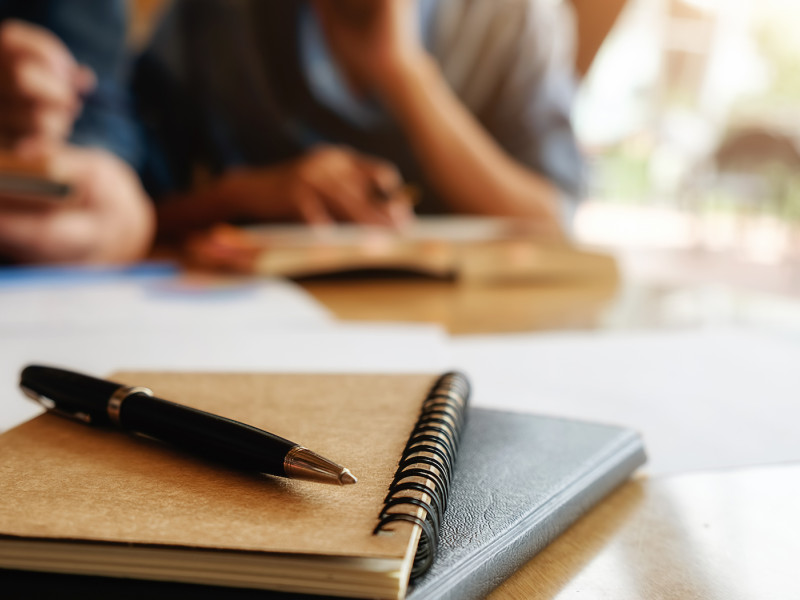 Op de voorgrond een notebook met pen, op de achtergrond mensen die samenwerken