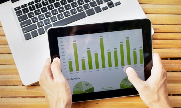 Twee handen met tablet met daarop een groene grafiek en laptop op achtergrond
