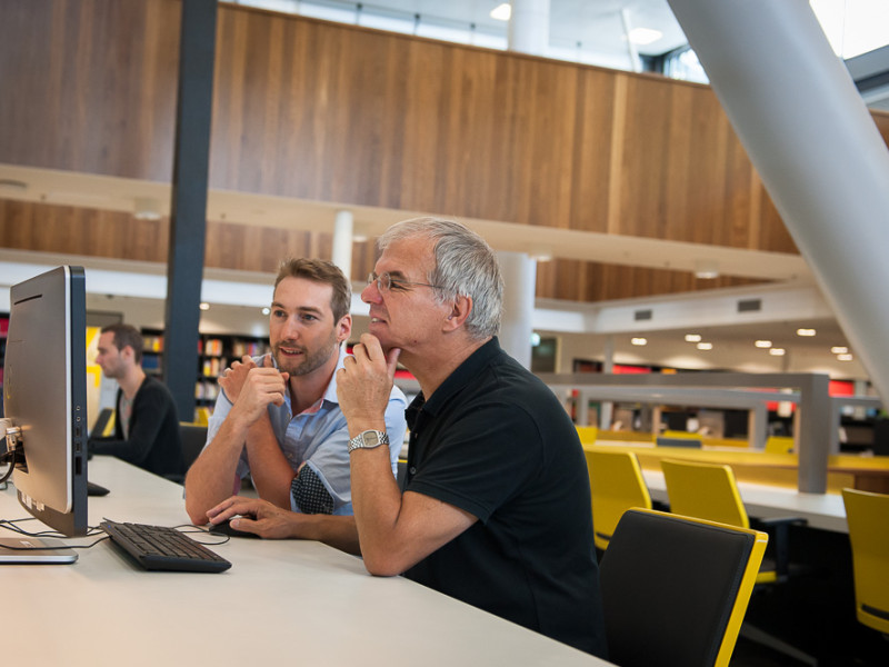 Oudere man met jonge man achter een computer in bibliotheek