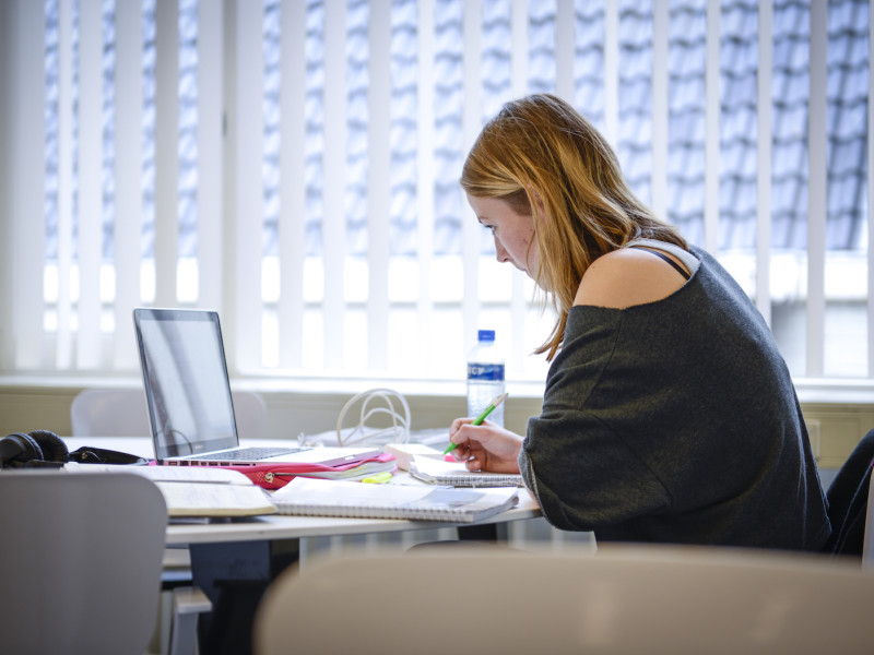 Studente aan het werk achter computer met veel documenten en aantekeningen voor zich