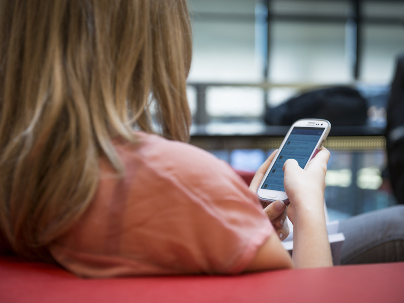 Vrouw van achter gefotografeerd zitten op een bank met een mobiele telefoon in haar hand