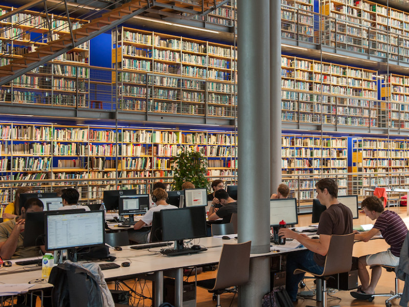 Open bibliotheek met hele hoge boekenwand en werkende studenten
