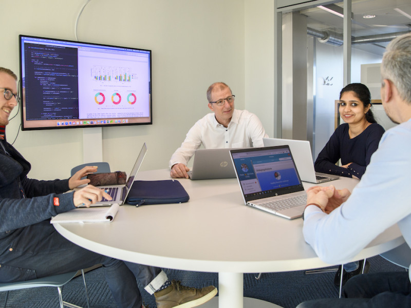 Vier mensen aan een ronde tafel met laptops en presentatiescherm