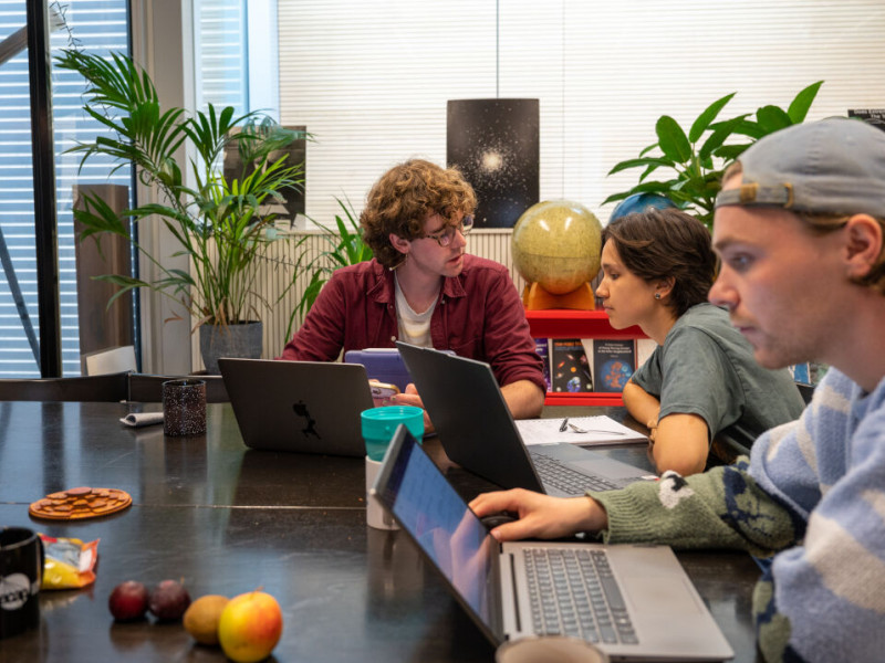 Drie studenten zitten aan een tafel, twee zijn in gesprek en de ander werkt op zijn laptop.