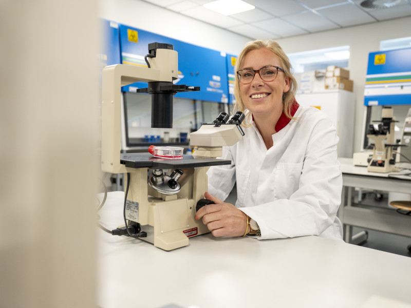 Anna Posthumus zit achter een microscoop in een lab, ze kijkt lachend de camera in. 