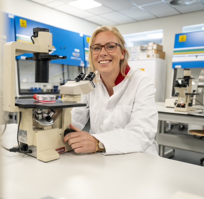 Anna Posthumus zit achter een microscoop in een lab, ze kijkt lachend de camera in. 