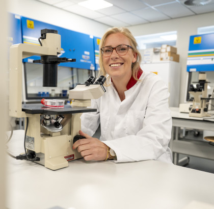 Anna Posthumus zit achter een microscoop in een lab, ze kijkt lachend de camera in. 